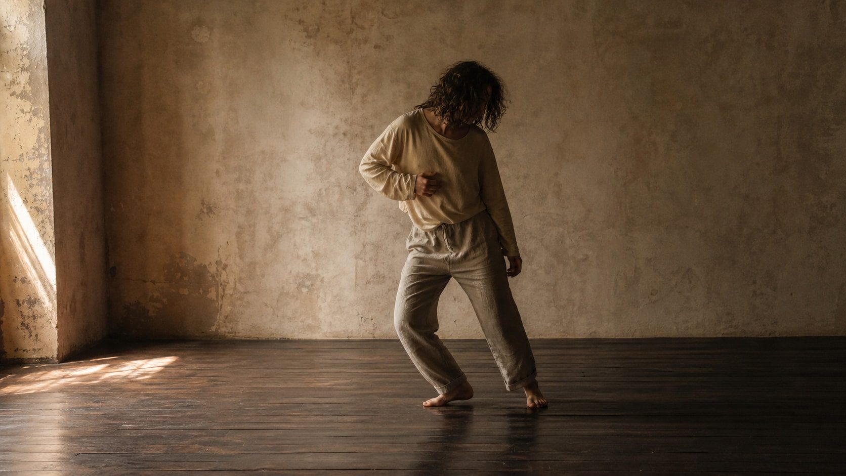 A barefoot dancer shifts through a small asymmetrical movement in a warm, timeworn room.